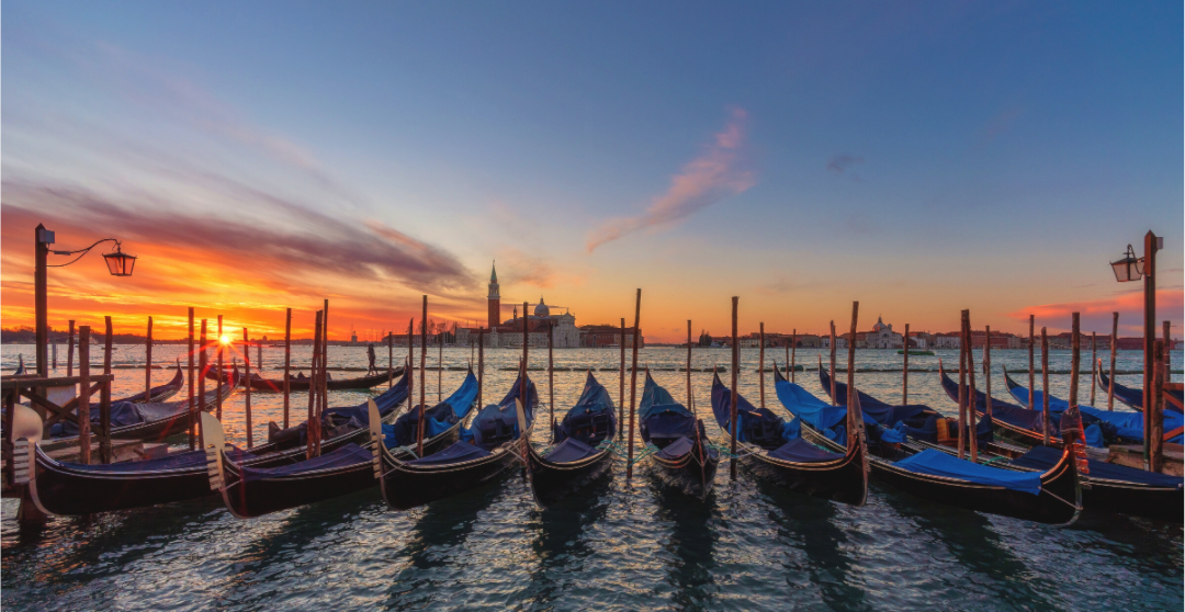 Gondels in Venetië bij zonsondergang met blauwe afdekzeilen, houten palen, San Marco-klokkentoren en warme oranje-roze lucht – romantische reisfotografie wanddecoratie.